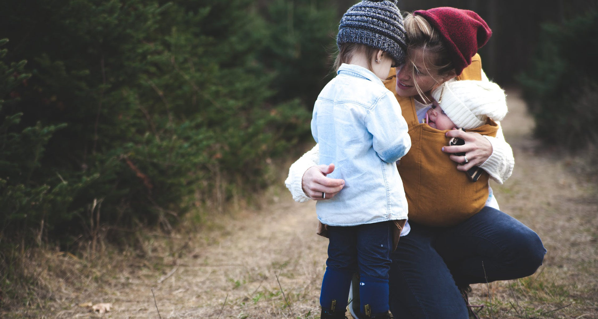 Outdoor photo of a mother kneeled down with a baby in a carrier and talking to a toddler