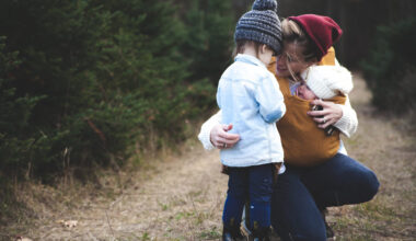 Outdoor photo of a mother kneeled down with a baby in a carrier and talking to a toddler