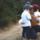 Outdoor photo of a mother kneeled down with a baby in a carrier and talking to a toddler